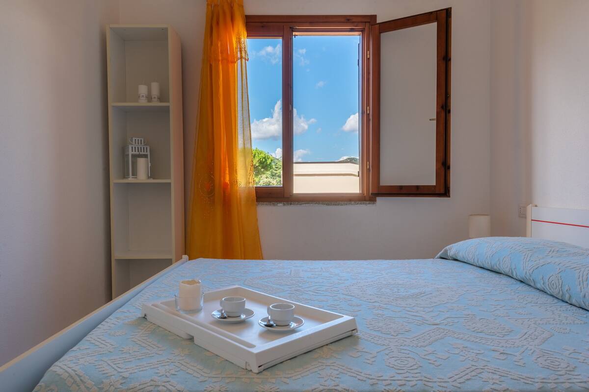 Une chambre avec un lit, une fenêtre donnant sur la plage, une étagère blanche avec quelques objets et un plateau avec des tasses et une bougie.
