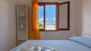 A bedroom with a bed, a window with a view of the beach, a white shelf with items, and a tray with cups and a candle.