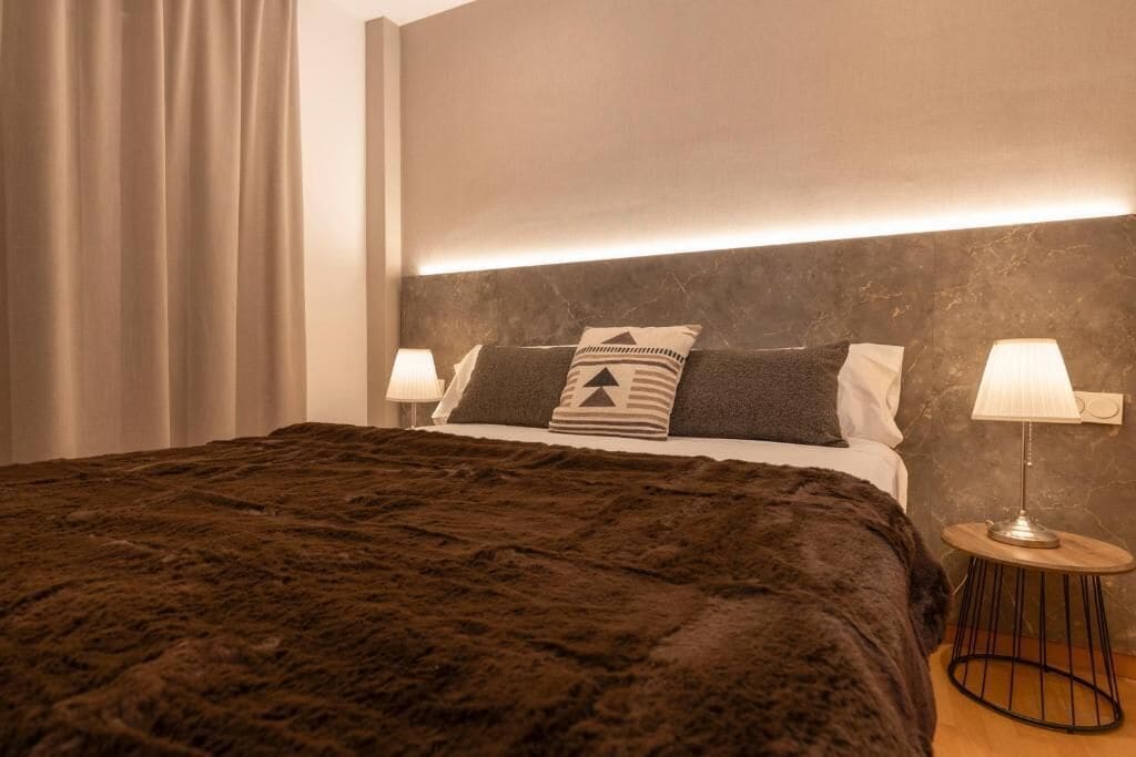 A modern bedroom with a large bed, a headboard with integrated lighting, two bedside lamps, and a dark brown shaggy rug.