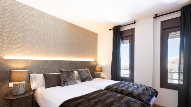 Un lit double avec une couvre-lit blanche et deux oreillers, une lampe de chevet et une vue sur l’extérieur par une fenêtre avec des rideaux.