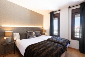 Cama doble con colcha blanca y dos almohadas, una lámpara de noche y vistas al exterior a través de una ventana con cortinas.