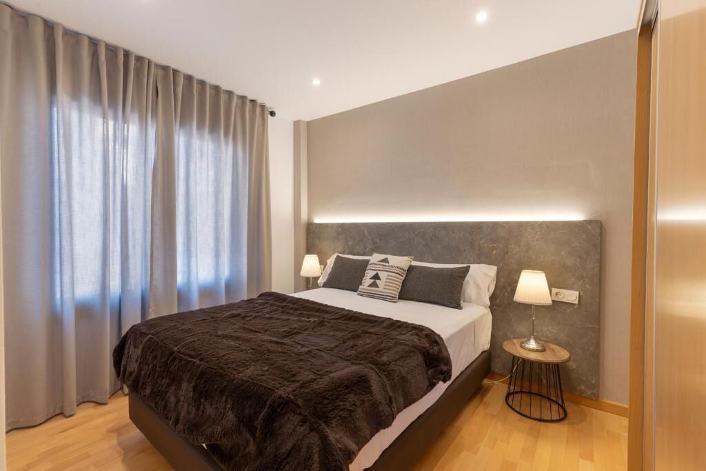 A modern bedroom with a large bed, two bedside lamps, and a view of the outdoors through sheer curtains.