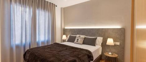 A modern bedroom with a large bed, two bedside lamps, and a view of the outdoors through sheer curtains.