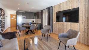 A modern living room with a wooden accent wall, a flat-screen TV, grey upholstered chairs, a coffee table with candles, and a view into the kitchen area.