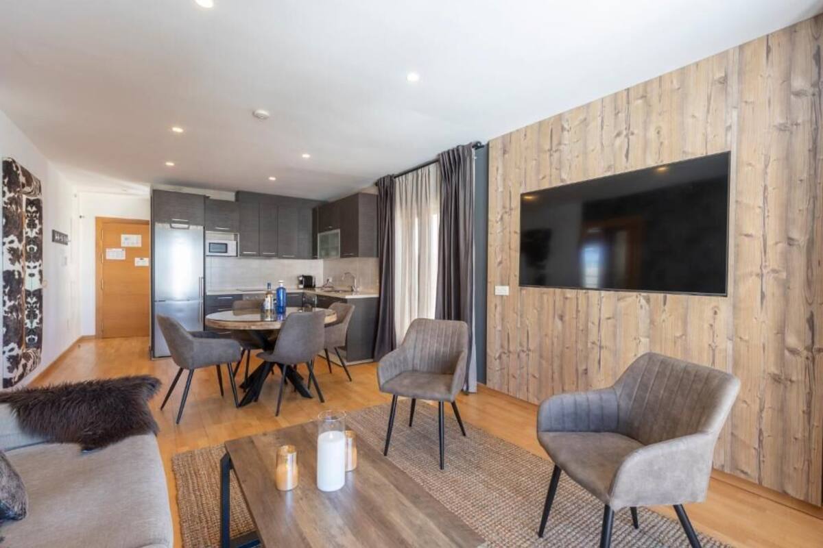Een moderne woonkamer met een houten accentmuur, een flatscreen televisie, grijs gestoffeerde fauteuils, een salontafel met kaarsen, en uitzicht op de keuken.