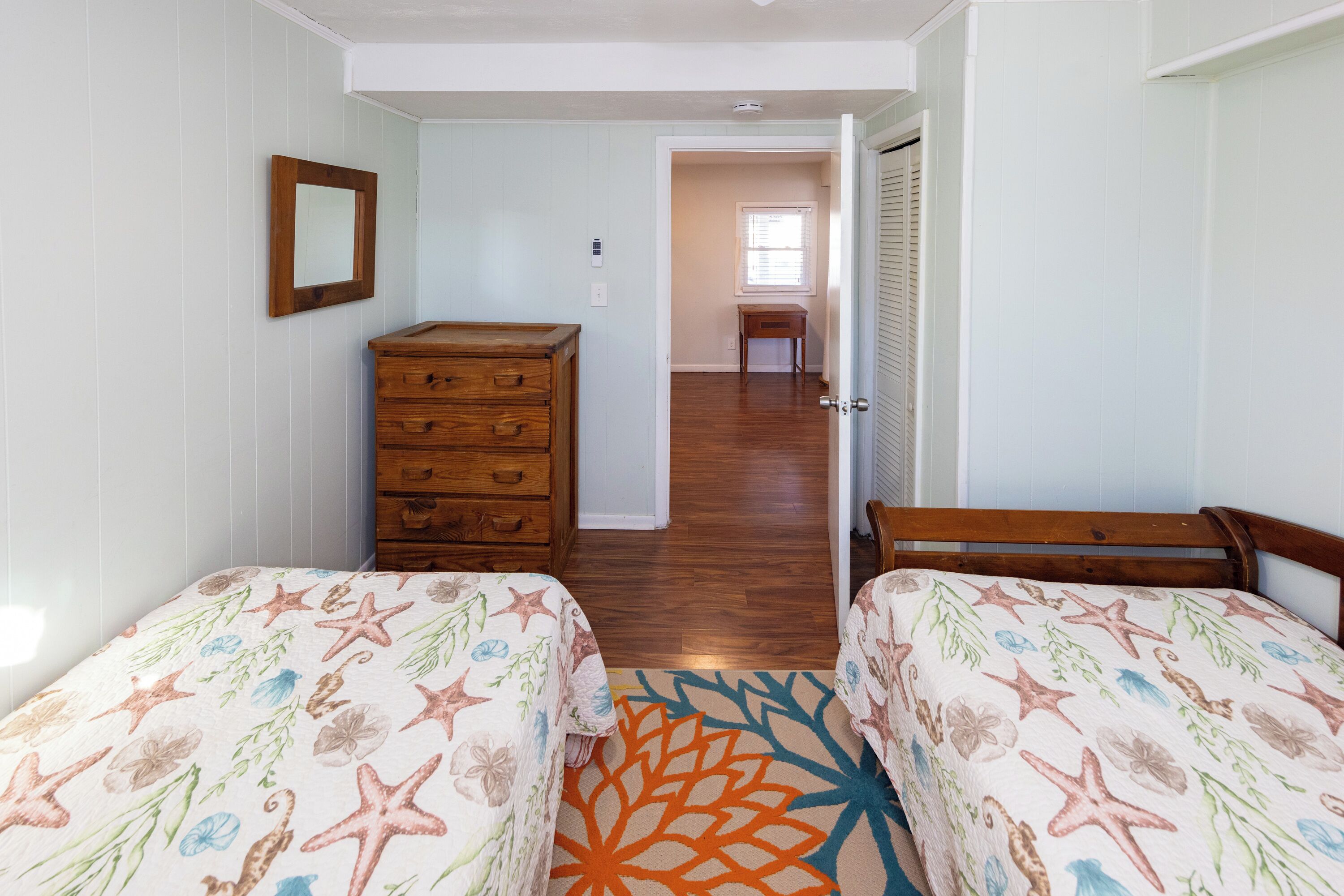 Une chambre avec deux lits, une commode et un miroir. Un couloir mène à une autre pièce.