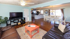 A living room with a brown leather sofa, a small orange coffee table, a flat-screen TV, and a kitchen area in the background.