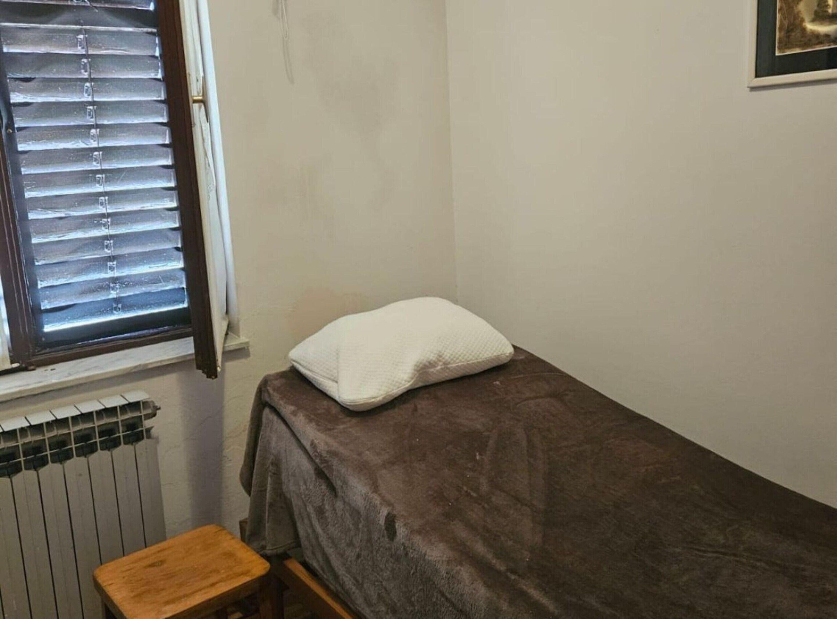 Ein kleines Zimmer mit einem Einzelbett, einem Fenster, einem Heizkörper und einem hölzernen Hocker.