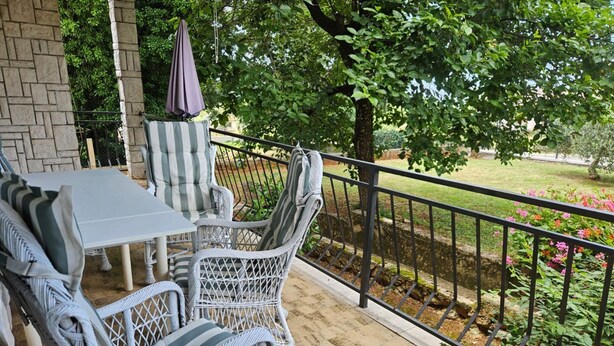 A covered patio with wicker furniture, a table, and chairs, overlooking a garden with flowers and a tree. - Apartment in Nature (Kastelir-Labinci)