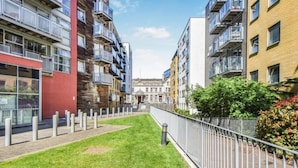 Une rue étroite en milieu urbain, bordée d’immeubles d’appartements modernes, avec une bande centrale enherbée et une clôture en métal.