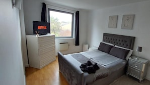 Une chambre à coucher comprenant un lit, une commode, un téléviseur diffusant Netflix, une fenêtre et deux œuvres d’art murales.