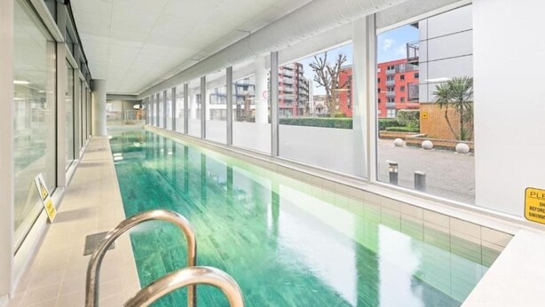 Une longue piscine intérieure aux parois en verre transparent, dotée d’une échelle en métal et offrant une vue sur les bâtiments de la ville.