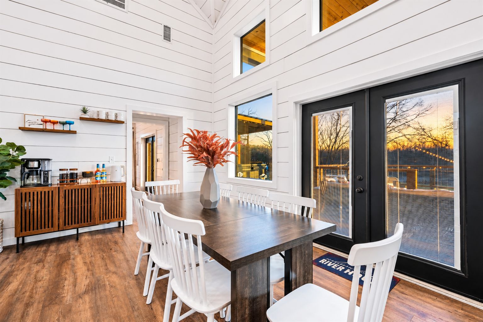 Un comedor con mesa de madera y sillas blancas, un área de cocina con mostrador de madera, y ventanas amplias que ofrecen vistas al exterior.