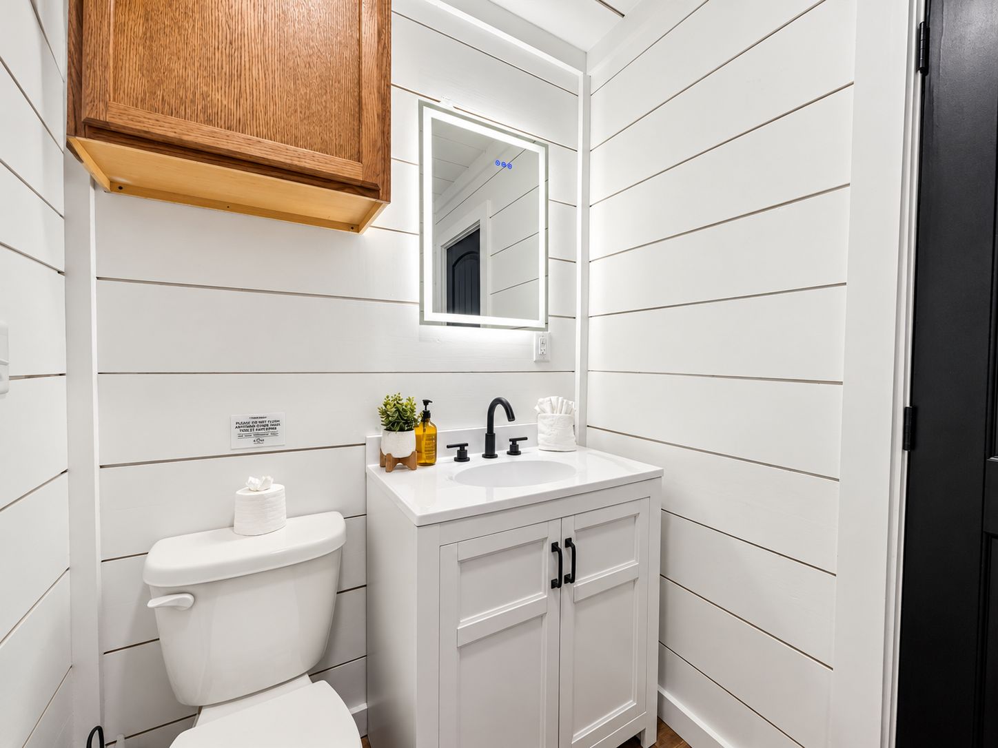 A bathroom with a white toilet, a wooden cabinet, a sink with a mirror above it, and a black door.