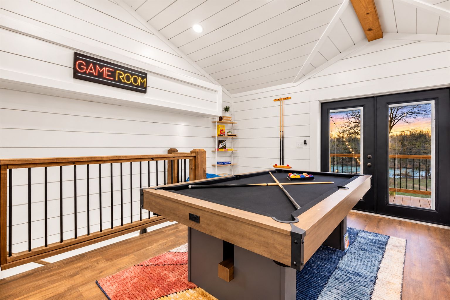 Une table de billard dans une pièce où est affiché un panneau indiquant « SALLE DE JEUX », avec des rampes en bois et une vue à travers une porte vitrée.