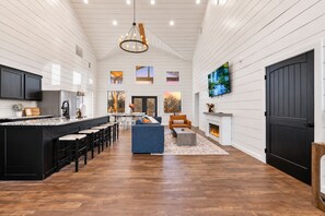 A spacious interior with a kitchen, dining area, and living room, featuring a fireplace and a large TV. - Cozy River Getaway W/ Heated Pool, Pool Table, Kayaks, Fishing | EV (Broken Bow)