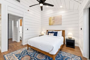 Queen Suite | Airy bedroom with white bedding, a ceiling fan, warm wood furniture, and blue accents. A calm space to recharge after hiking or exploring Broken Bow.