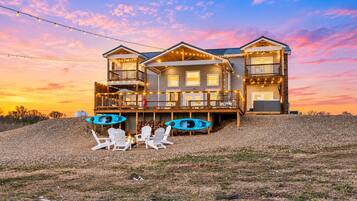 A two-story house with a deck, outdoor seating, kayaks, and a scenic sunset view.