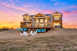 A two-story house with a deck, outdoor seating, kayaks, and a scenic sunset view.