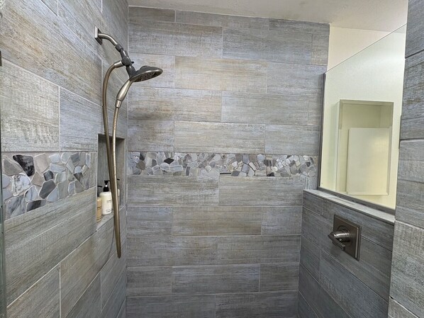 Ducha con cabezal de ducha manual, pared con azulejos y detalle de piedra, y puerta con manija.