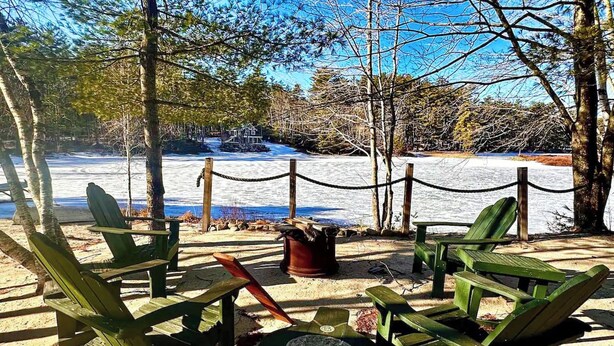 Un espace aménagé avec des chaises et des tables en bois, un foyer et une vue sur un paysage enneigé, avec des arbres et une étendue d’eau.