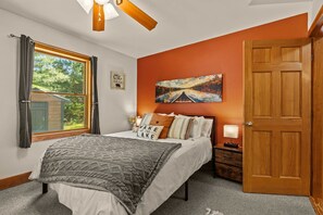 A bedroom with a bed, a nightstand, a lamp, a window with curtains, a painting on the wall, and a door.