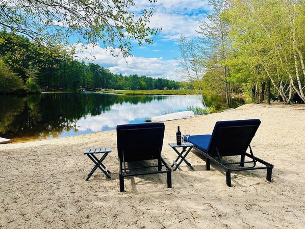 A lakeside setting with two lounge chairs and a small table, a bottle of wine, and a clear view of the lake and surrounding trees.