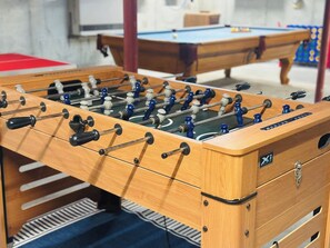 A wooden foosball table with a drawer and a logo, set in a room with a pool table in the background.
