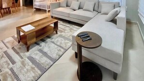 A modern living room with a sectional sofa, a coffee table, and a side table. There is a ceiling fan, an air conditioning unit, and a window with blinds. - New and stylish 3-Bedroom Apartment (Dar es Salaam)