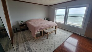 A bedroom with a bed, a nightstand, a lamp, a window with a view of the ocean, and a patterned rug. - 3 BR Lake Front Getaway! Dog Friendly, Lakeside Deck, Dock, Incredible Sunrises (Alpena)