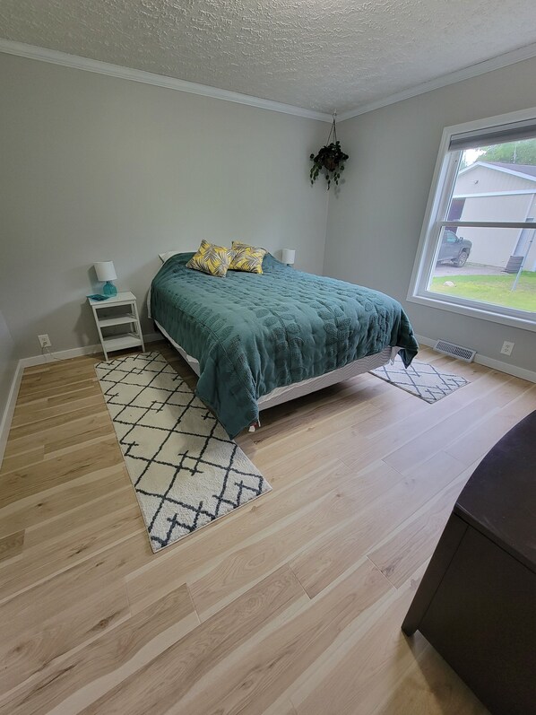 A bedroom with a bed, a nightstand, a hanging plant, a window with a view of a vehicle, and a patterned rug. - 3 BR Lake Front Getaway! Dog Friendly, Lakeside Deck, Dock, Incredible Sunrises (Alpena)