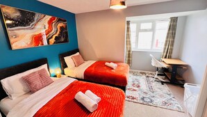 A hotel room with two beds, a desk, a chair, a large abstract painting, and a view of the city through the window.