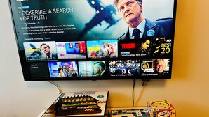 A television displaying a drama titled "LOCKERBIE: A SEARCH FOR TRUTH" with a subtitle about Colin Firth. Below the TV, there are two game boxes labeled "GAME HOUSE" and "SCRABBLE".