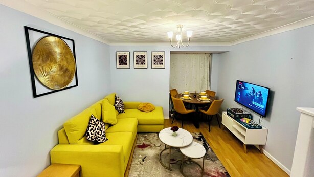A living room with a yellow sofa, a dining table with chairs, a TV, and framed artwork on the wall.