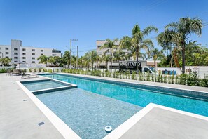En swimmingpool med kristallklart blått vatten, omgiven av palmer och en byggnad med skylten ”HABITAT SOFAS”.