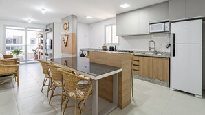 Una cocina moderna con una isla central, muebles blancos y un microondas. Hay una zona de comedor con sillas de mimbre y una mesa de vidrio.