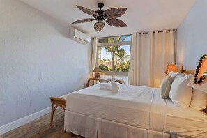 Une chambre d’hôtel avec un lit, un ventilateur de plafond, une fenêtre avec des rideaux et une vue sur des palmiers.