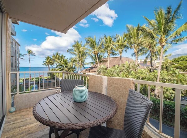 Un balcon avec une table ronde, des chaises en osier et une vue sur les palmiers et l’océan.