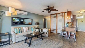 A living room with a sofa, coffee table, and a TV. There's a kitchen area with a bar and stools. - Oceanview updated 1 bedroom condo, AC, 6 mile sandy beach (Kihei)