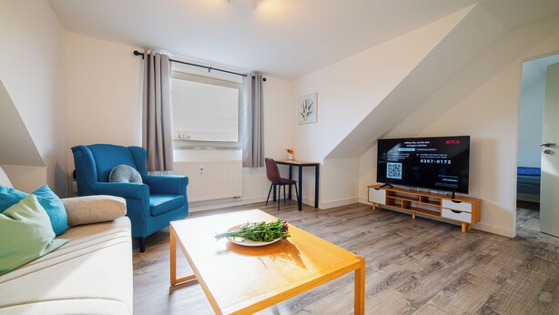 A modern living room with a sofa, a blue armchair, a wooden coffee table with a flower arrangement, a TV on a wooden stand, and a dining area with a table and chairs. - Central feel-good apartment 1 - ideal for your vacation or business trip (Magdeburg)