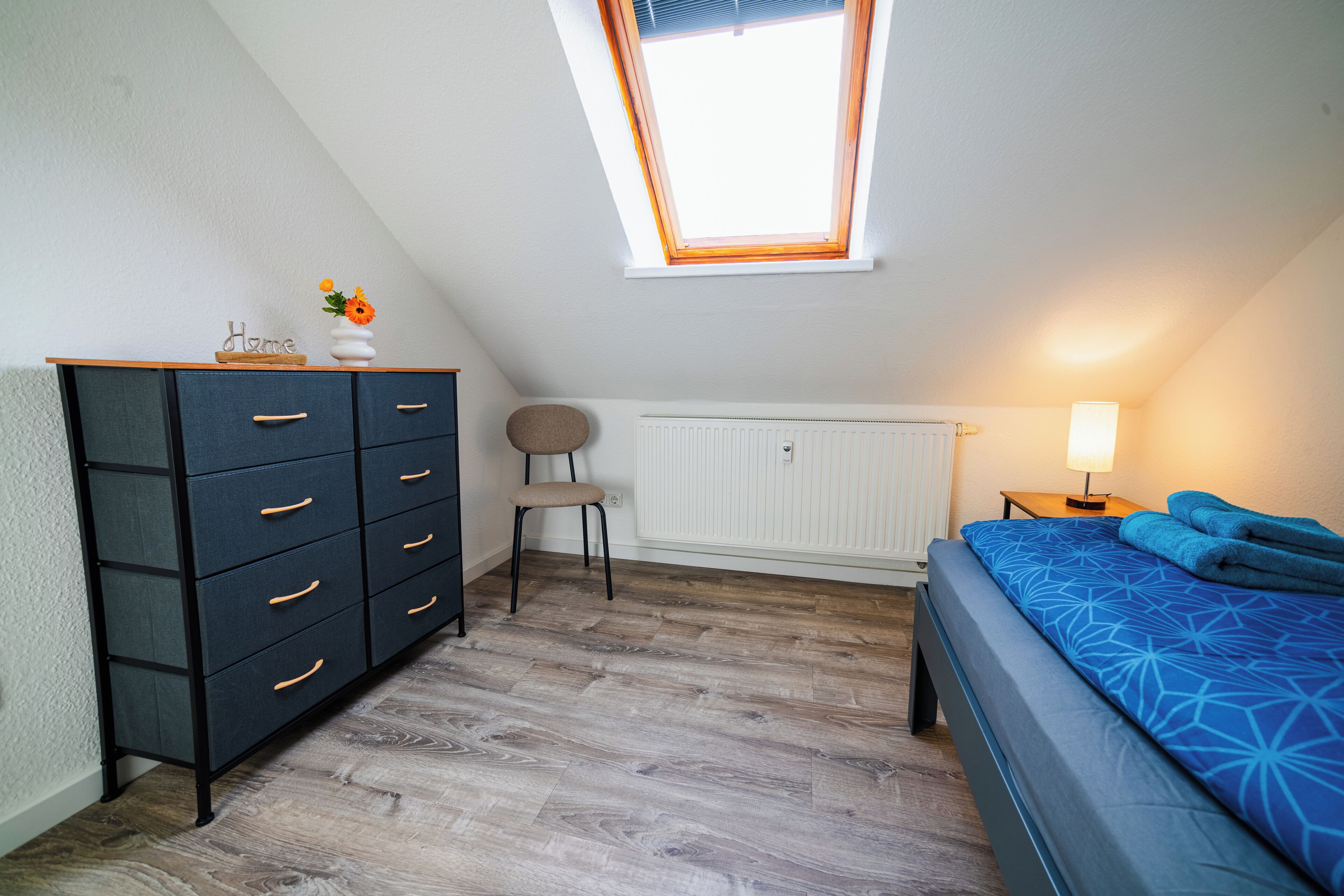 Ein Schlafzimmer mit Dachfenster, einem Schminktisch, einem Sessel, einer Heizung, einem Nachttisch mit Lampe und einem Bett mit gemustertem Bettbezug.