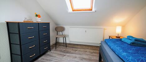 Ein Schlafzimmer mit Dachfenster, einem Schminktisch, einem Sessel, einer Heizung, einem Nachttisch mit Lampe und einem Bett mit gemustertem Bettbezug.