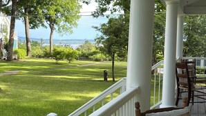 A porch with a white railing, a wooden chair, and a view of a grassy yard with trees and a distant body of water. - Charming 5-bedroom Roaring Brook cottage in Harbor Springs with beach access. (Harbor Springs)