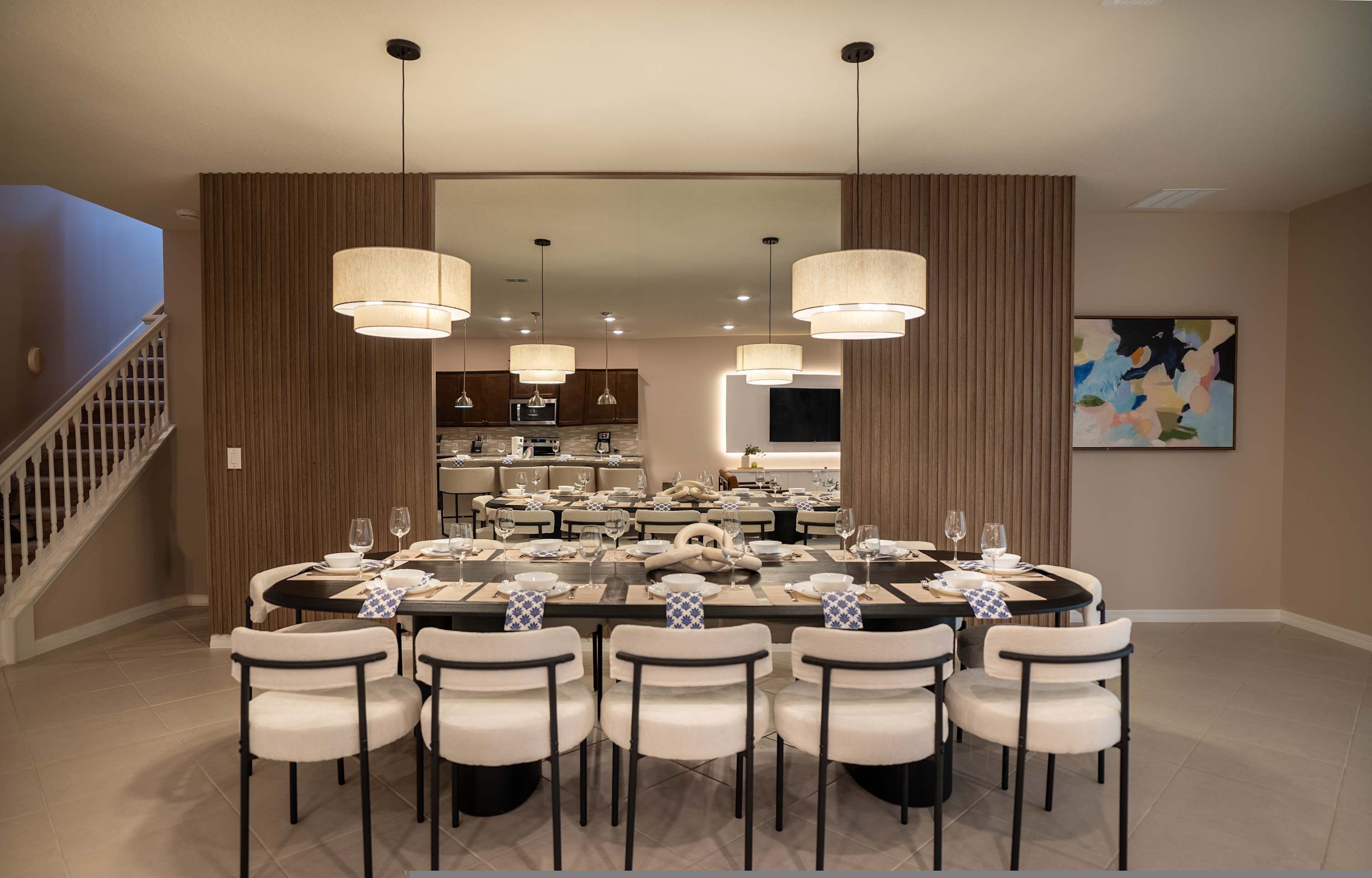A dining area with a large round table set for eight, surrounded by white chairs. There are pendant lights above the table, and a kitchen is visible in the background.