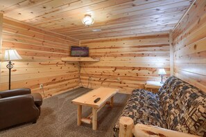 A cozy cabin interior with wooden walls, a sofa, a coffee table, and a TV mounted on the wall. - Waccabuck | 17.5 Private Acres, Pool, Hot Tub (Logan)