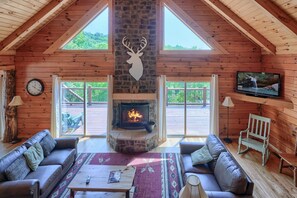A cozy cabin interior with a stone fireplace, wooden walls, two sofas, a television, and large windows. - Waccabuck | 17.5 Private Acres, Pool, Hot Tub (Logan)