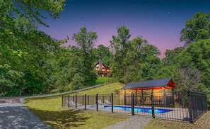 A lodge with a swimming pool, surrounded by a fence and trees, at dusk.