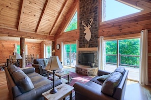 A log cabin interior with a stone fireplace, leather sofas, and large windows offering a view of the outdoors.