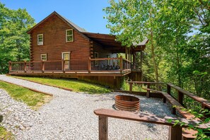 A rustic log cabin with a deck, surrounded by gravel and greenery.