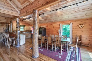 A spacious cabin interior with wooden floors, a dining area with wooden chairs, a kitchen with wooden cabinets, and a large window providing a view of greenery. - Waccabuck | 17.5 Private Acres, Pool, Hot Tub (Logan)